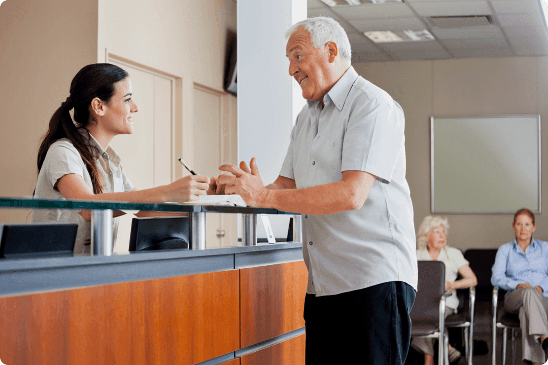 An elderly man speaks to a smiling receptionist at a desk, with three people seated in the waiting area behind them.